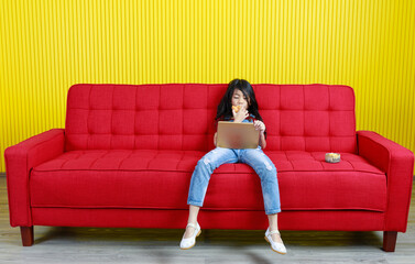 Studio shot of Asian young primary school girl in fashionable denim jeans overalls outfit sitting crossed legs on red couch browsing surfing internet with touchscreen tablet computer in living room
