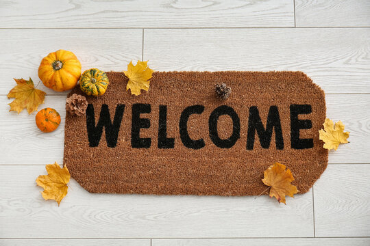 Door mat with word WELCOME, fallen leaves and pumpkins on white wooden floor