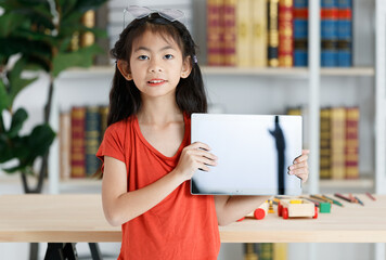 Portrait studio shot Asian clever smart young primary school girl daughter in casual outfit holding black blank screen tablet computer in hands smiling look at camera in front bookshelf background