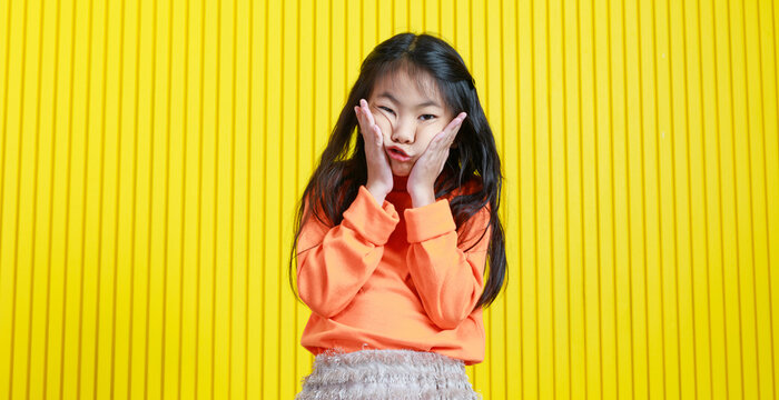 Portrait studio shot Asian playful cheerful funny face young primary school girl daughter in casual outfit holding hands squeezing cheeks and mouth looking at camera on yellow stripe wall background