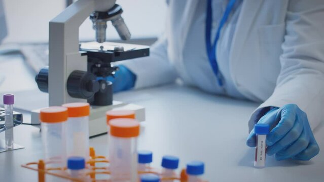 Close up of scientist wearing white coat looking through microscope and making notes whilst holding blood sample in test tube which he puts on desk to show label type ab - shot in slow motion