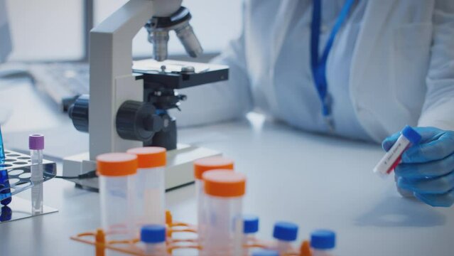 Close up of scientist wearing white coat looking through microscope and making notes whilst holding blood sample in test tube which he puts on desk to show label type b - shot in slow motion