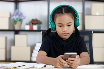 Portrait shot of Asian young cute happy primary schoolgirl in casual outfit sitting at working table smiling wearing headphone listening to music song playlist from touchscreen smartphone in hands