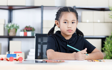Asian young smart clever elementary schoolgirl artist in casual outfit sitting using color pencils drawing painting picture on paper alone on working table with tablet and wooden toy in living room