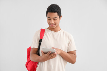 Male African-American student with tablet computer and backpack on light background