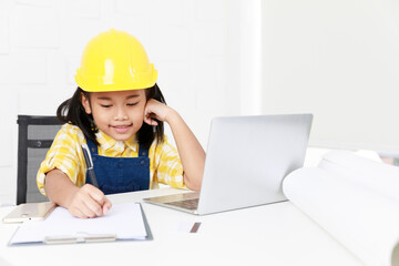 Asian young little girl dreaming about future job career as engineering wearing yellow safety hardhat helmet sitting on working table with laptop computer blueprint smartphone and paper clipboard