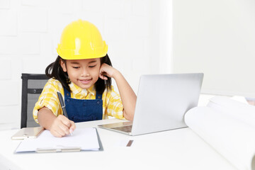 Asian young little girl dreaming about future job career as engineering wearing yellow safety hardhat helmet sitting on working table with laptop computer blueprint smartphone and paper clipboard