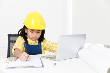 Asian young little girl dreaming about future job career as engineering wearing yellow safety hardhat helmet sitting on working table with laptop computer blueprint smartphone and paper clipboard