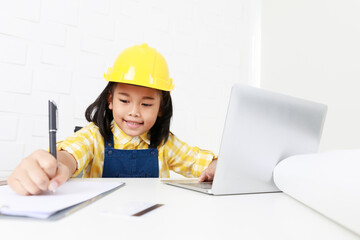 Asian young little girl dreaming about future job career as engineering wearing yellow safety hardhat helmet sitting on working table with laptop computer blueprint smartphone and paper clipboard