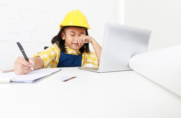 Asian young little girl dreaming about future job career as engineering wearing yellow safety hardhat helmet sitting on working table with laptop computer blueprint smartphone and paper clipboard