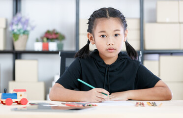 Asian young smart clever elementary schoolgirl artist in casual outfit sitting using color pencils drawing painting picture on paper alone on working table with tablet and wooden toy in living room