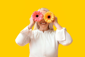 Happy mature woman closing face with gerbera flowers on yellow background. International Women's Day celebration