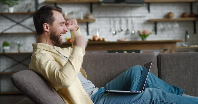 Happy young man celebrate success win online bet and showing YES gesture. Lucky male freelancer reading great news online using laptop while resting on cozy sofa at home.