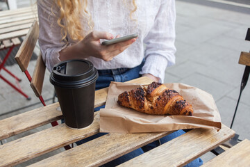 Young white blond woman having a break and holds smartphone in hand. Street veranda table in cafe with cardboard paper cup of coffee to go and french croissant, . business breakfast concept