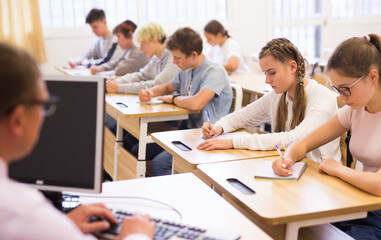 Fototapeta premium Focused teenage students studying in classroom with teacher, writing lectures in workbooks