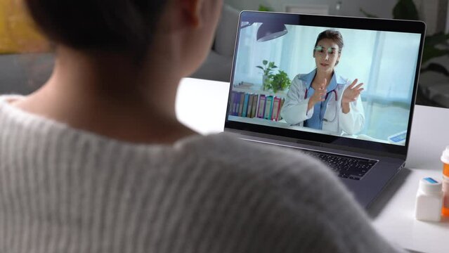 Young Asian woman sit on couch at home have video call with doctor use wireless internet connection on laptop, female patient talk consult with physician online on webcam conference on computer