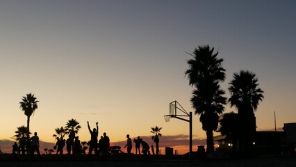 Silhouettes of players on basketball court outdoor, people playing basket ball game, sunset ocean beach, California coast, USA. Black hoop, net and backboard on streetball sport field. Mission beach.