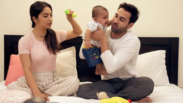 A young couple struggling with their upset crying son while sitting on the bed. An Indian young man and his wife playing with their sweet toddler in their leisure time - family bonding  happy child...