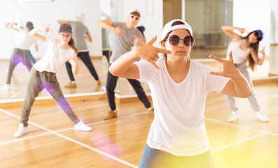Fototapeta premium Teenage girl hip hop dancer in casual clothes, cap and sunglasses exercising with friends at dance center