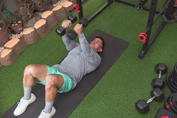 A fit asian man in a gray sweater does floor dumbbell floor flys on a black mat. Chest workout at...