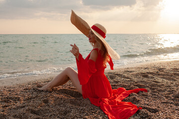 Beautiful strong woman wearing maxi straw hat and red chiffon cape like dresss walking in the beach...