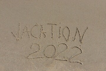 The inscription vacation 2022 on the sand by the water and the rising wave, seashore beach vacation by the sea