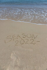 The inscription season 2022 on the sand by the water and the rising wave, seashore beach vacation by the sea