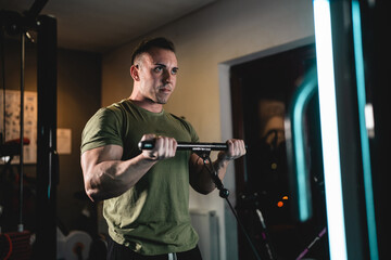 One man young adult caucasian male bodybuilder training arms bicep on the cable machine in the gym holding bar wearing shirt dark photo real people copy space side view