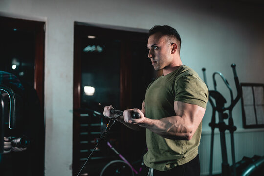 One man young adult caucasian male bodybuilder training arms bicep on the cable machine in the gym holding bar wearing shirt dark photo real people copy space side view