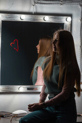 Stylish beautiful young woman with hairstyle in fashionable clothes sits near a vintage mirror with lamps in the dark. Heart of red lipstick on the mirror