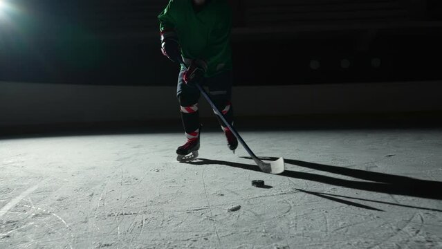 Low angle hockey forward player masterfully dribbles, hitting puck with stick scores goal. Hockey puck hits the net. An athlete man plays hockey on dark ice arena with spotlights. Slow motion.