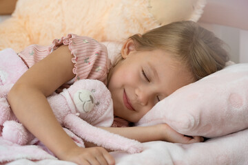Little girl sleeps at home in bed with toy and smiles. Child in dream hugs teddy bear. Orthopedic pillow, mattress and healthy sleep concept