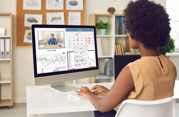 Busy young Afro American woman working at office desk with desktop computer, chatting with coworker, organizing schedule in calendar planner, having conference call with colleague. View over shoulder