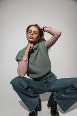 Stylish beautiful young girl model in fashionable vintage clothes with jeans, sweater and shirt with glasses sitting and posing in the studio with hard light