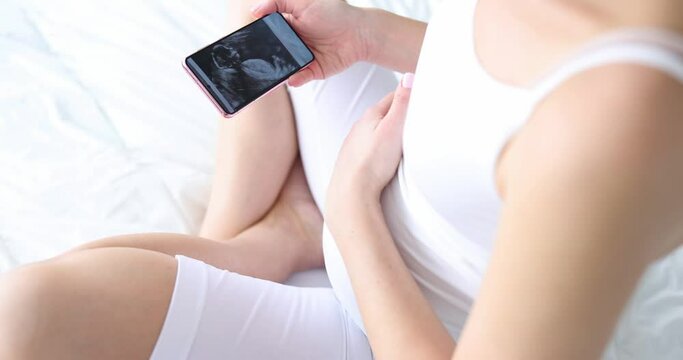 Pregnant woman sitting on bed and looking at ultrasound picture on mobile phone closeup 4k movie slow motion