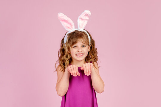 Adorable funny little kid with curly blond hair in dress and bunny ears, smiling and showing paws gesture while imitating rabbit during Easter party in pink studio
