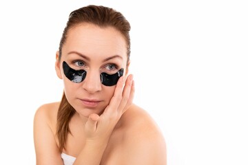 Fototapeta premium a woman applies cosmetic black patches under her eyes against a white wall