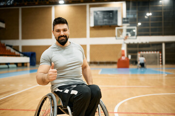 Happy wheelchair-bound athlete in wheelchair shows thumb up on basketball court and looks at camera.