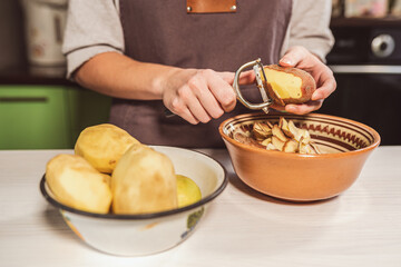 Young woman housewife in brown chef's apron peels potatoes with kitchen paring knife on blurred background of kitchen interior