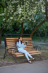 A teenage girl in denim clothes is talking on the phone, sitting on a bench in a city park. Spring. Apple trees in bloom. 