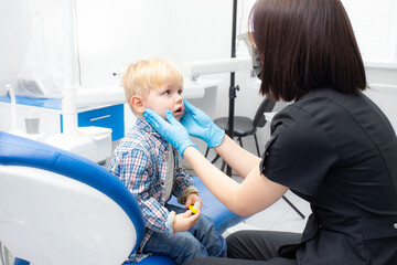 A cute little boy sits at the dentist, opened his mouth, showing his teeth to the dentist. Happy boy is sitting in the dentist's chair. Dentist - young brunette woman