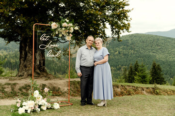 an elderly wedding couple stands near a stylish wedding arch under a tree against the backdrop of...