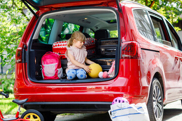 Cute little preschool girl sitting in car trunk before leaving for summer vacation with parents....