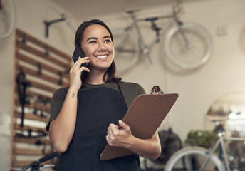 You can come through to collect your bike. Shot of an attractive young woman standing alone in her bicycle shop and using her cellphone while holding a clipboard.