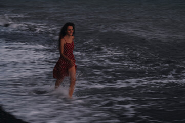 A happy young girl in a dress runs along the sea waves on the beach on a summer evening.