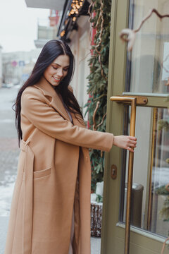 Young Brunette Woman Wearing Beige Coat Opening Green Glass Door, Entering To Cafe. Street Style Fashion