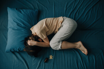 Top view of a young woman in bed with pills lying next to her. An unrecognizable woman is depressed or experiencing mental problems.