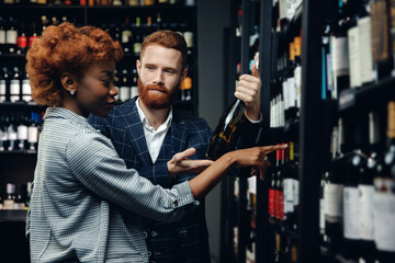 Fototapeta na wymiar Lover multicultural couple talking and choosing wine in shop. Man and African girl are buying alcohol