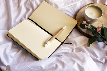 Blank notebook, candle and flowers on white sheet. Relaxing scene. Flat lay, top view. 