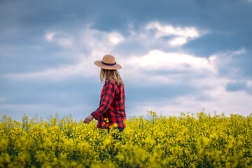 Fototapeta na wymiar Proud farmer is looking at blooming oilseed field. Woman with straw hat and plaid shirt standing in rapeseed agricultural field